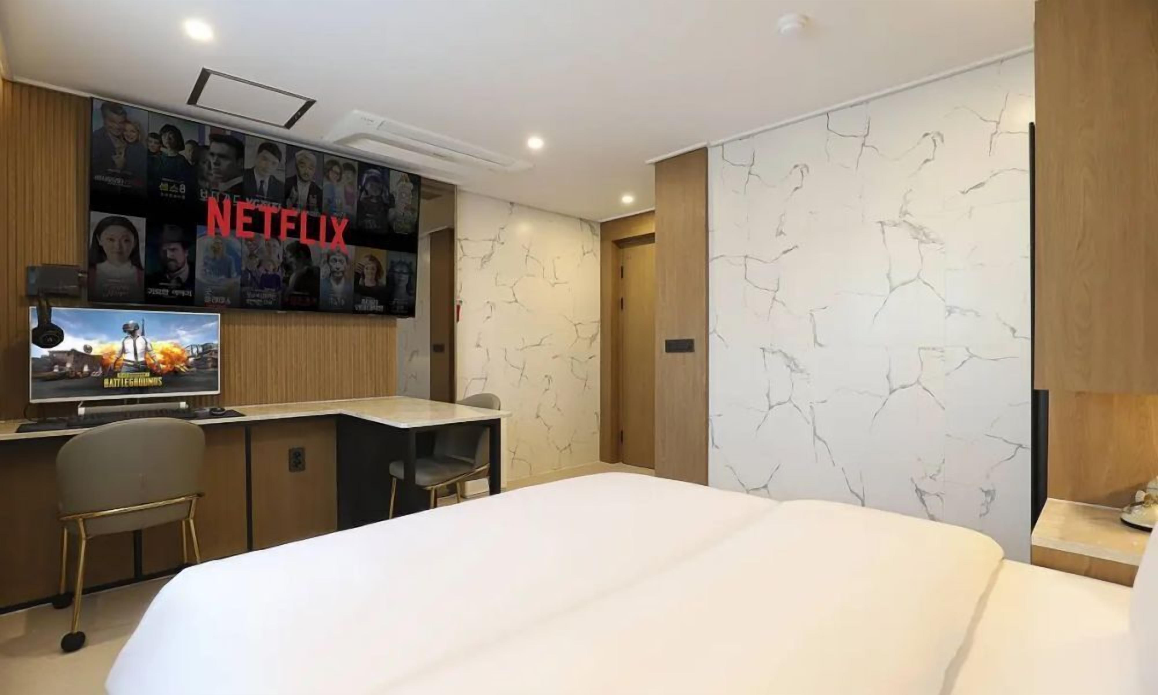 Ein Hotelzimmer mit Bett, Schreibtisch, Stuhl, einem an der Wand befestigten Fernseher und einer Tür.