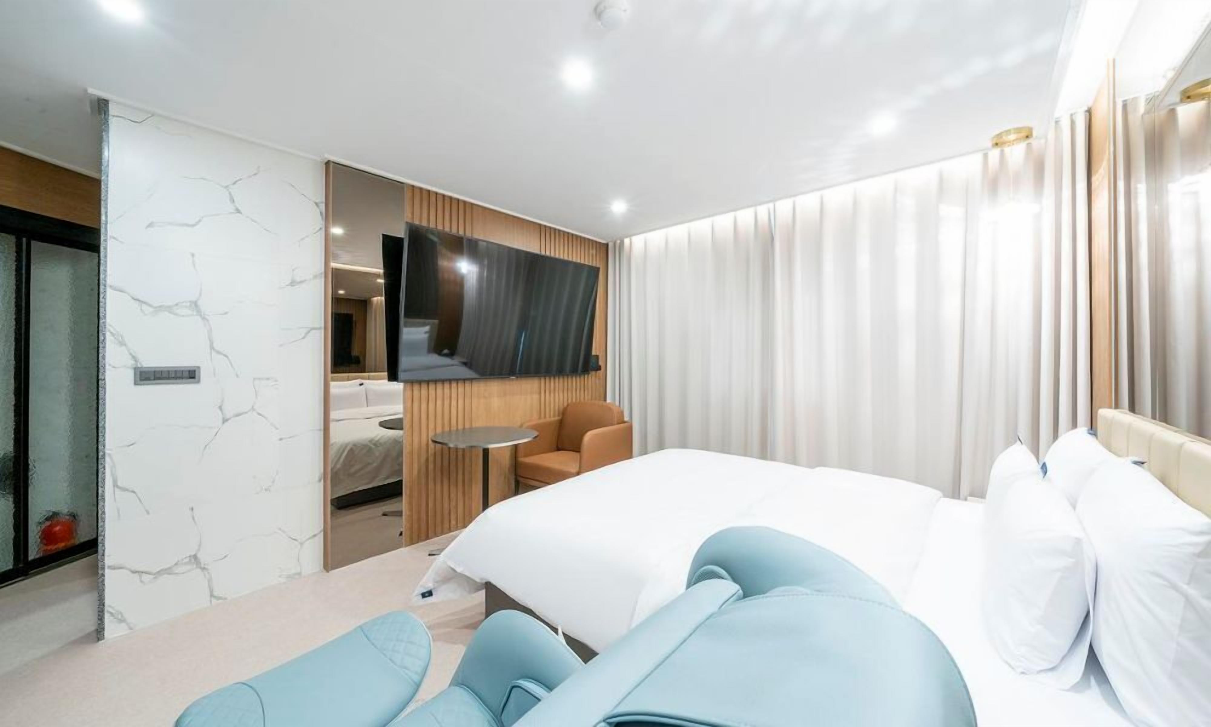 Una habitación de hotel moderna con una cama grande, un televisor montado en la pared, una zona de estar con una mesa y un baño visible a través de una puerta de cristal.
