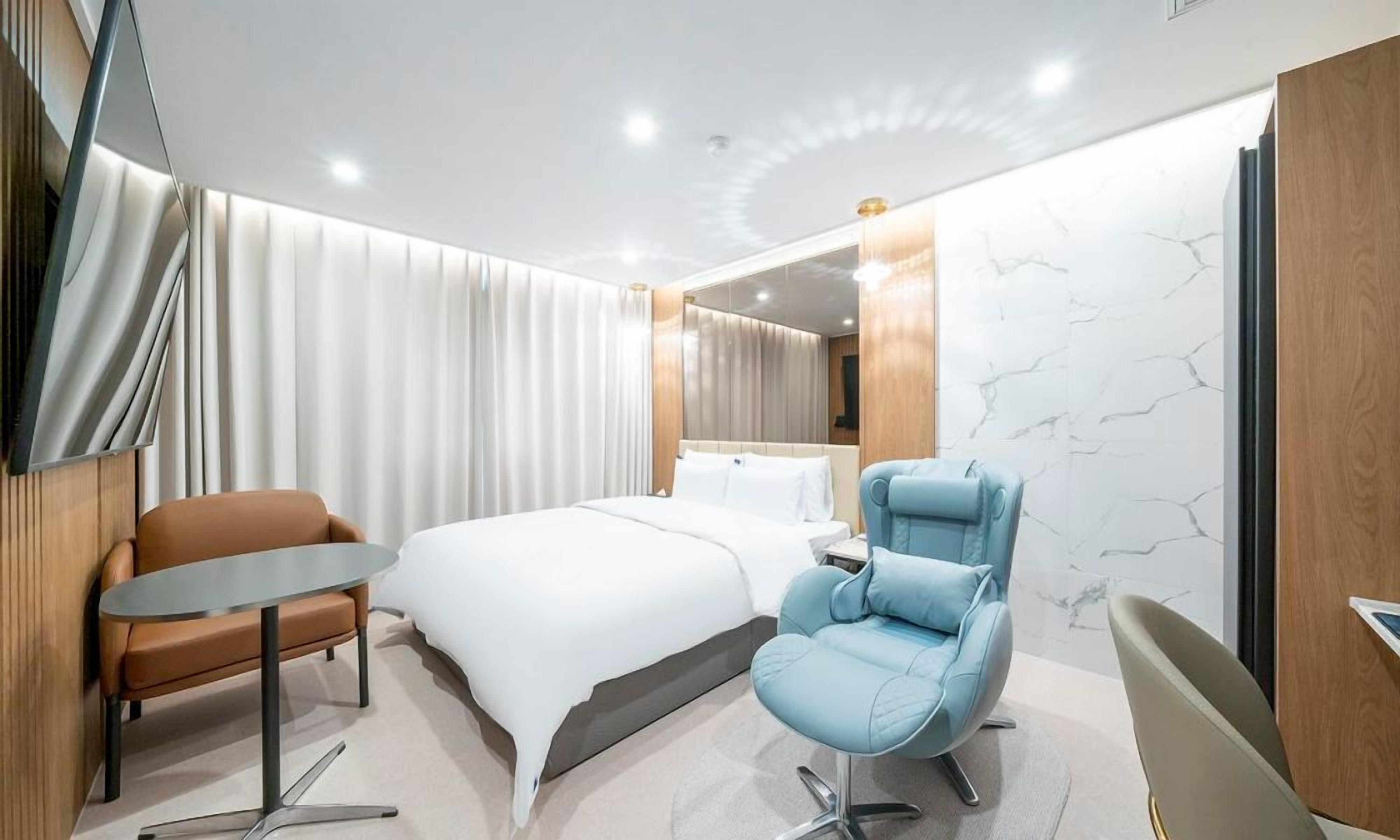 Una habitación de hotel moderna con una cama, un sillón azul, una mesita y un espejo.