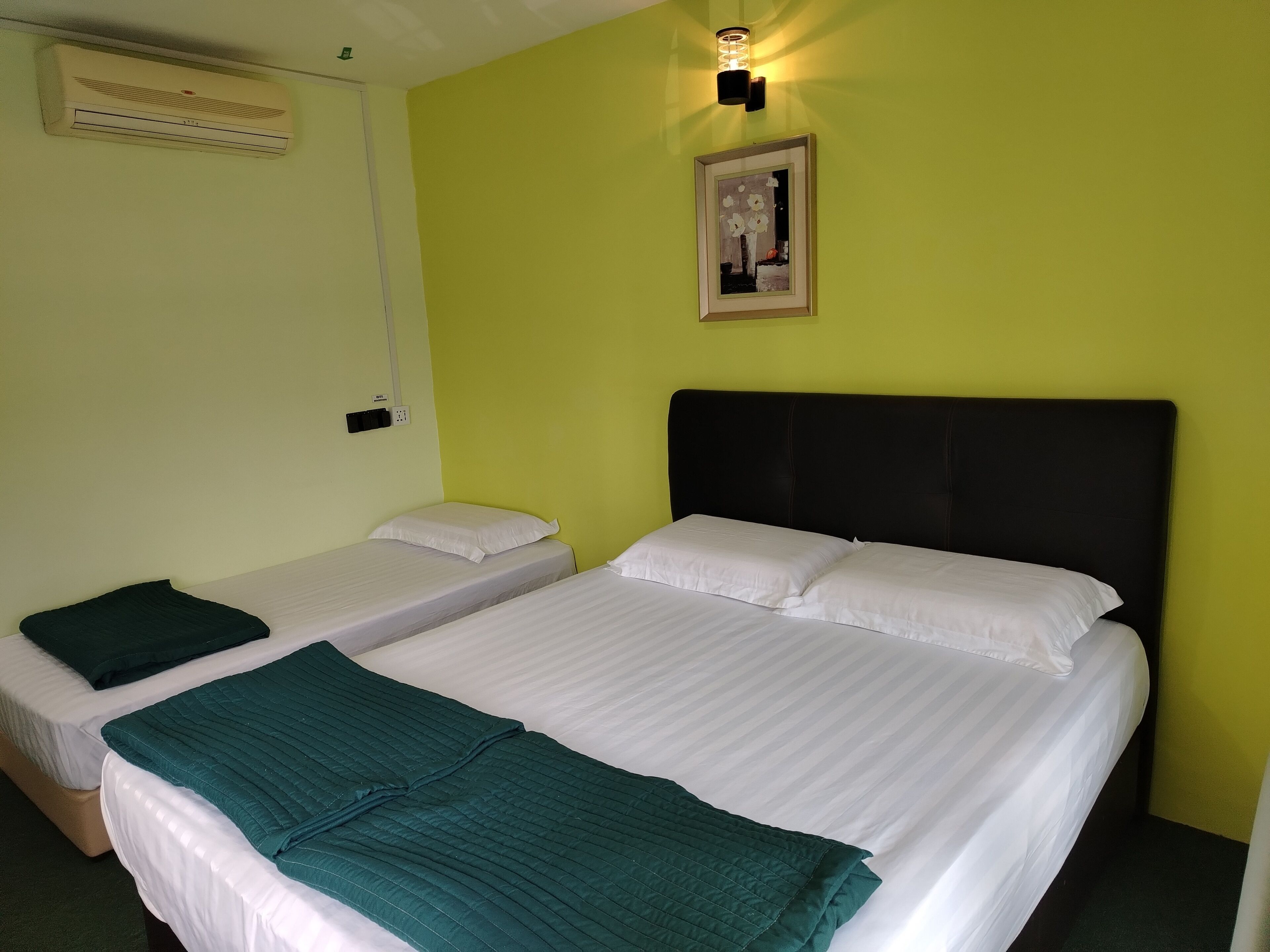 Une chambre d’hôtel avec deux lits simples, un grand lit, une couverture verte, un tableau encadré au mur et un ventilateur de plafond.