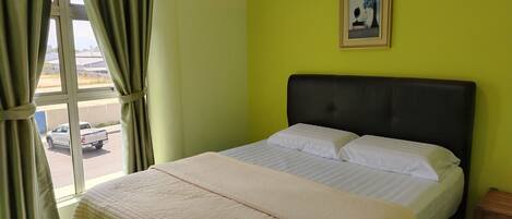 A hotel room with a bed, a window with curtains, a small wooden stool, and a wall-mounted picture.