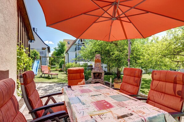 An outdoor patio with a table covered in a tablecloth, surrounded by chairs, and an umbrella providing shade.
