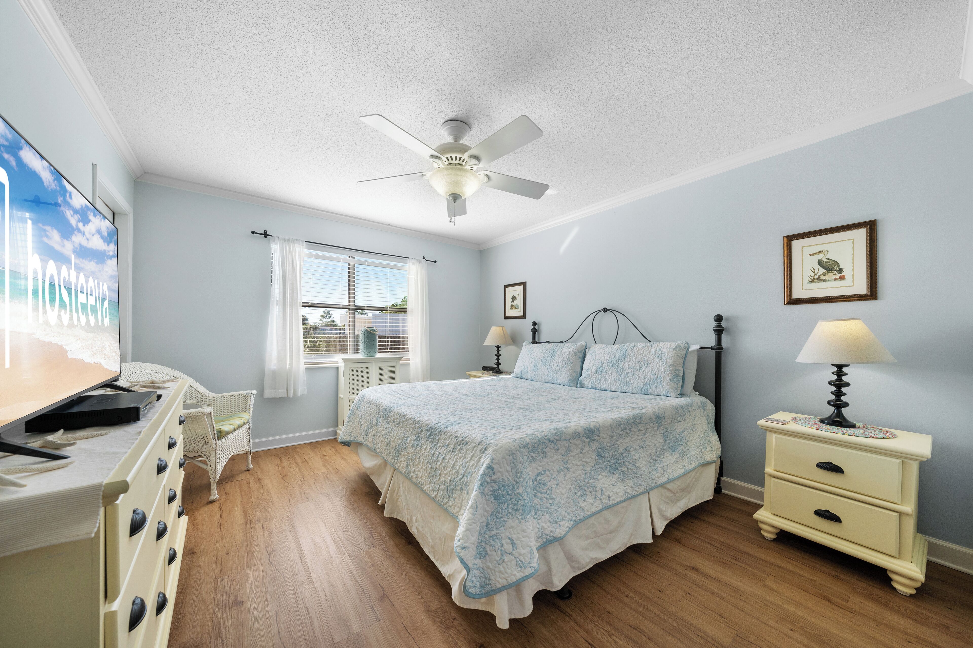 A bedroom with a bed, bedside tables, a ceiling fan, a window with blinds, and a wall-mounted TV.
