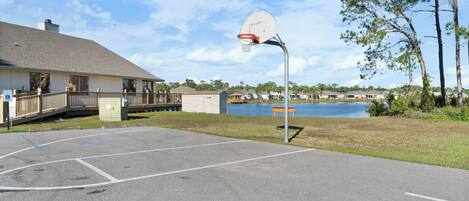 Un terrain de basket en plein air, avec un panier de basket, un banc et une vue sur un lac et des maisons en arrière-plan.
