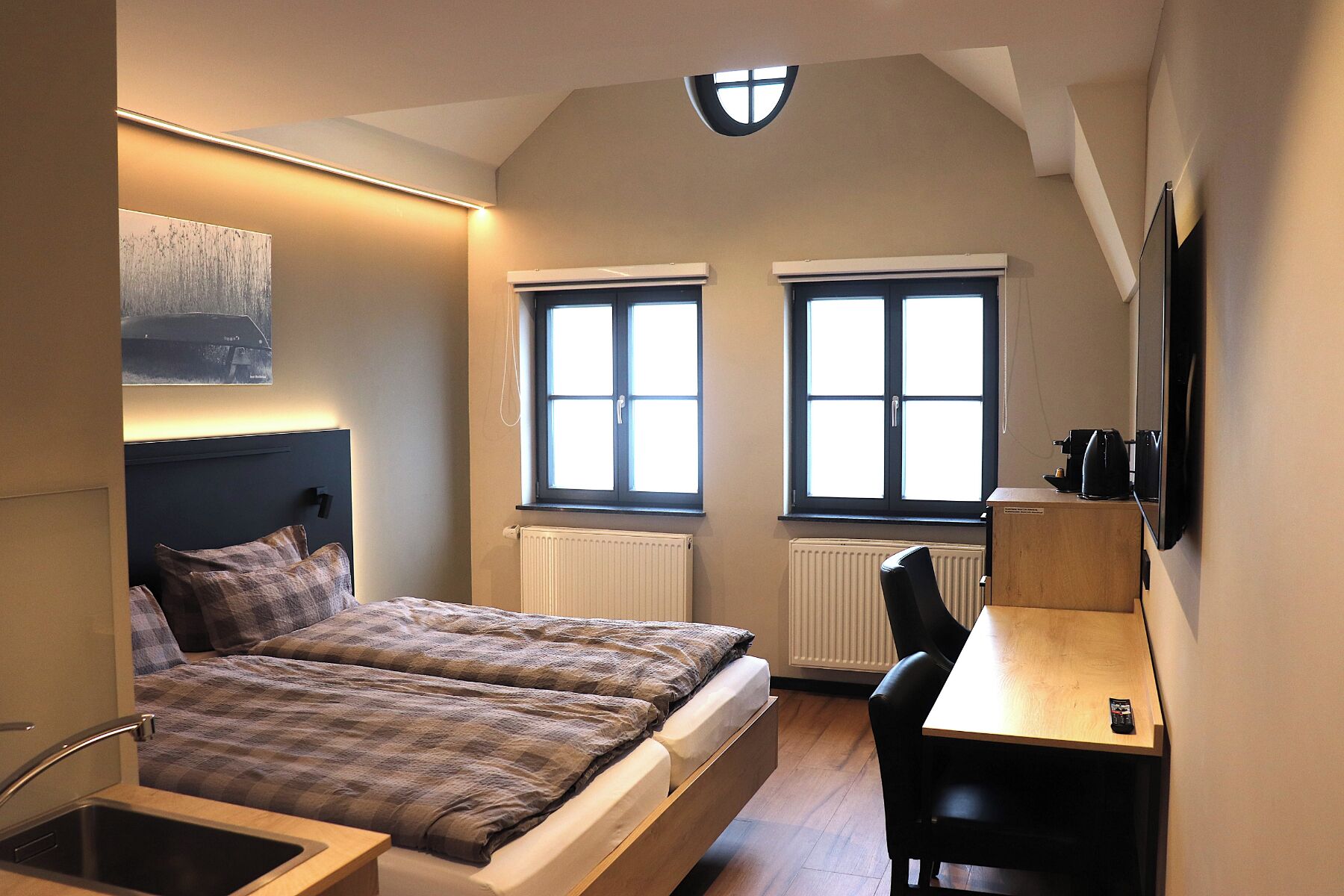 Quarto compacto com cama, escrivaninha e duas janelas.