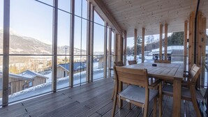 Una terraza de madera con una mesa y sillas, ventanas amplias y vistas a edificios nevados y montañas.