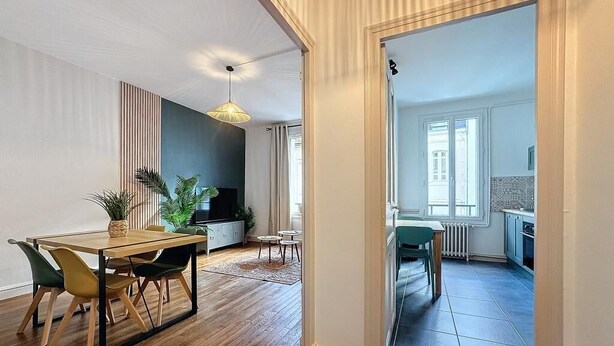 Un salon moderne avec une table à manger en bois, des chaises et un téléviseur. On aperçoit un coin cuisine à travers une porte ouverte.