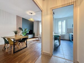 A modern living room with a wooden dining table, chairs, and a TV. There is a kitchen area visible through an open doorway. - Le 2-F2 historic center (MONTLUCON)