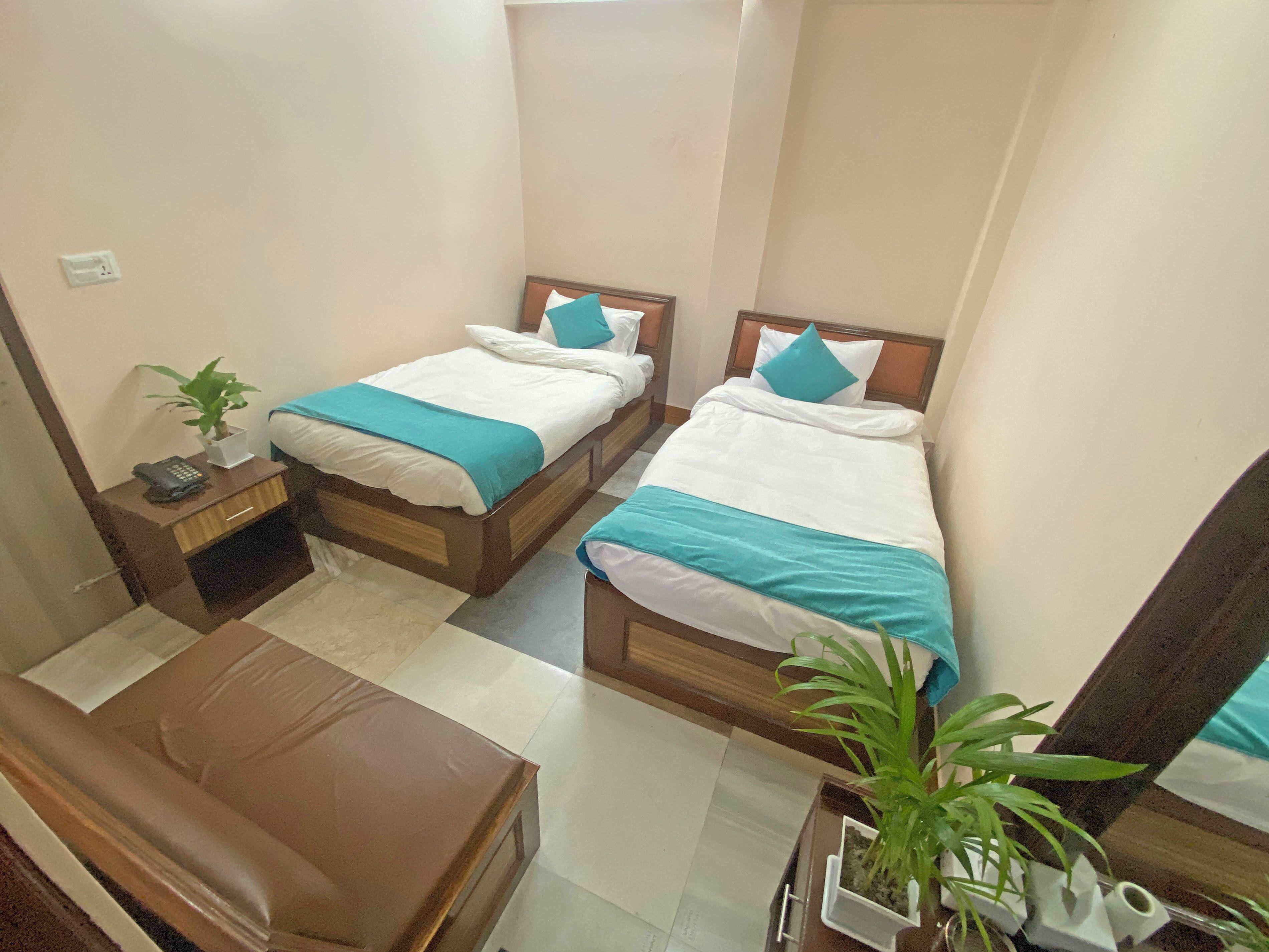 A small hotel room with two single beds, a wooden nightstand with a phone, a small table, and a chair. There is a potted plant on the nightstand and another on the floor.