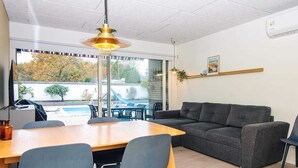 A modern living room with a wooden dining table, grey sofas, and chairs. A large window offers a view of a swimming pool and trees. - 4 Person Holiday Home in Glesborg (Glesborg)