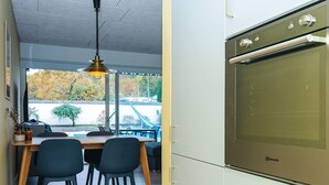 A modern kitchen with a built-in oven, a dining area with two chairs, and a view of a swimming pool and trees through large windows. - 4 Person Holiday Home in Glesborg (Glesborg)