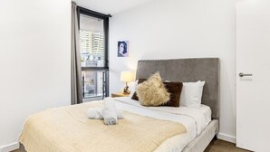 Una habitación de hotel con una cama, una ventana que ofrece vistas a edificios, una mesita de noche con una lámpara y un cuadro en la pared.