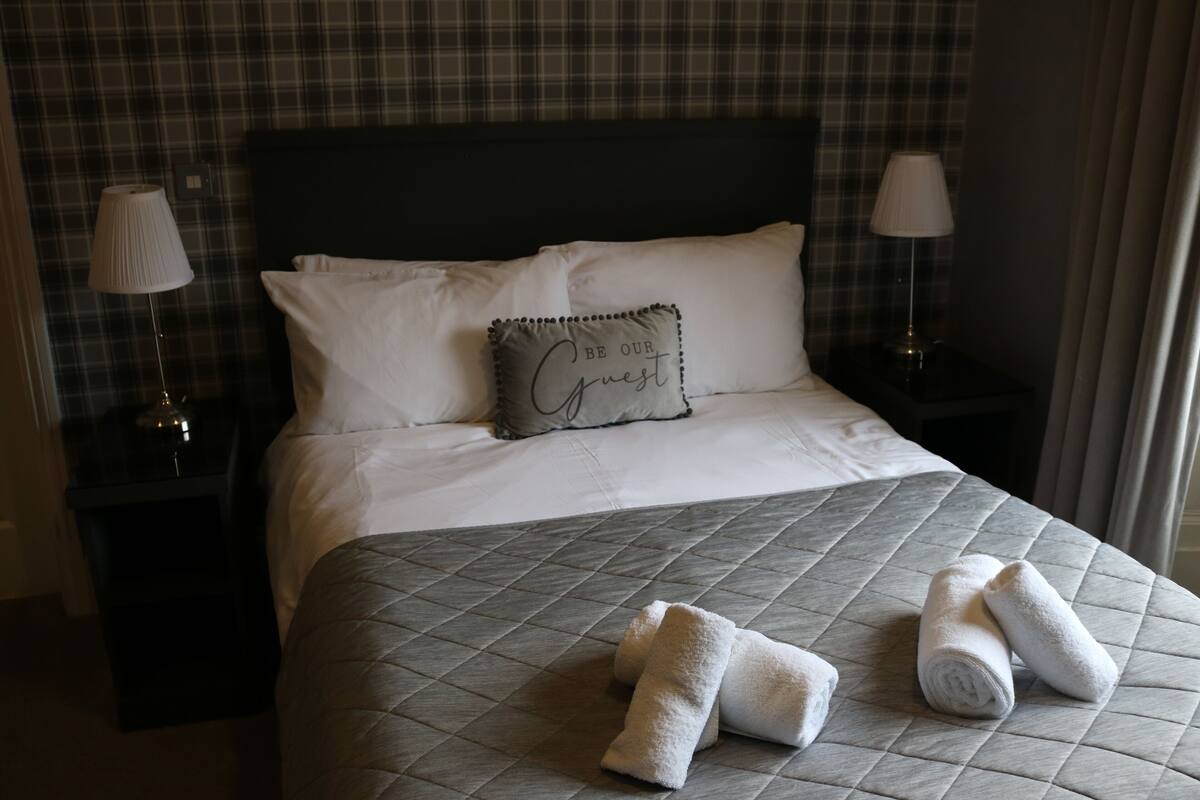 A hotel room with a bed, two bedside lamps, a pillow with text, and rolled towels on the bed.