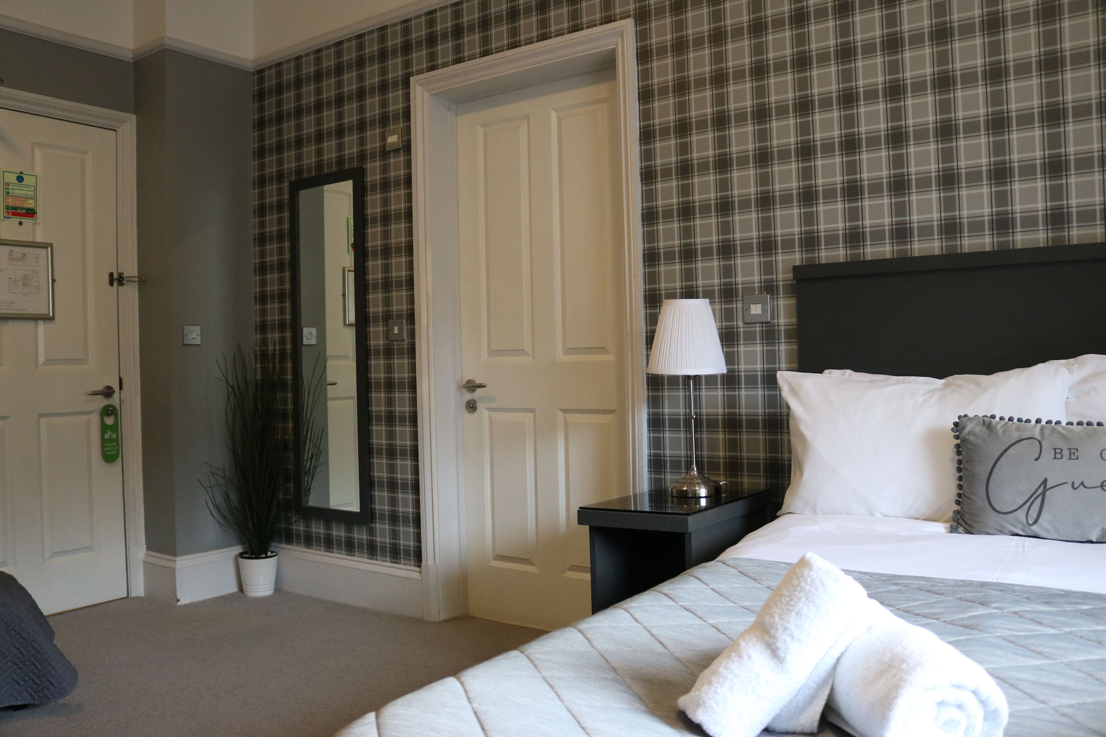 A hotel room with a bed, a nightstand, a lamp, a mirror, and a door.