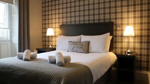 A neatly made bed with a quilted bedspread, two pillows, and rolled towels. There are two bedside lamps on black nightstands. The room has plaid wallpaper and a window with curtains. - Norfolk Arms Hotel (Glossop)