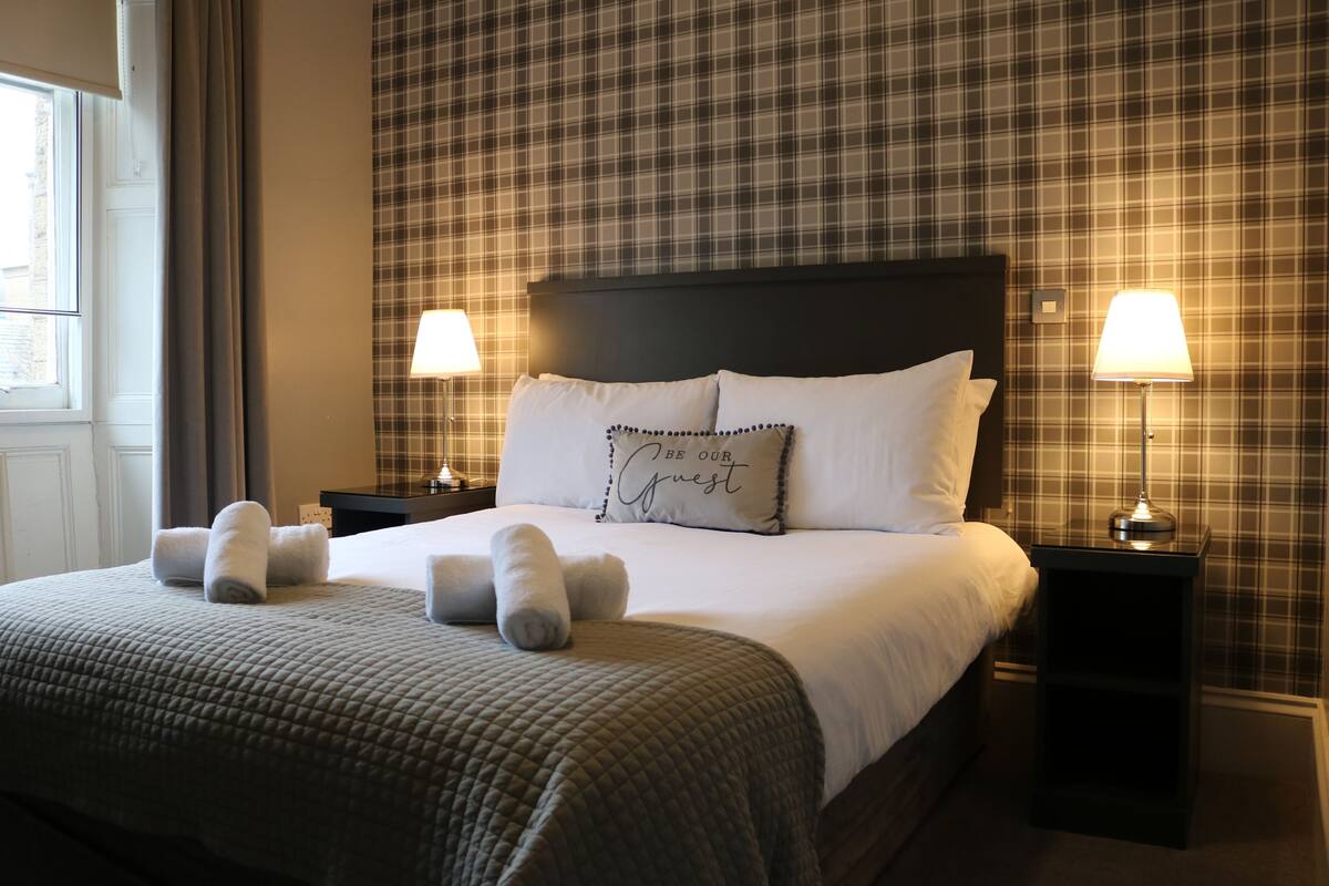 A neatly made bed with a quilted bedspread, two pillows, and rolled towels. There are two bedside lamps on black nightstands. The room has plaid wallpaper and a window with curtains.