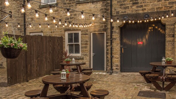 An outdoor patio area with wooden tables and benches, string lights, and potted plants. - Norfolk Arms Hotel (Glossop)