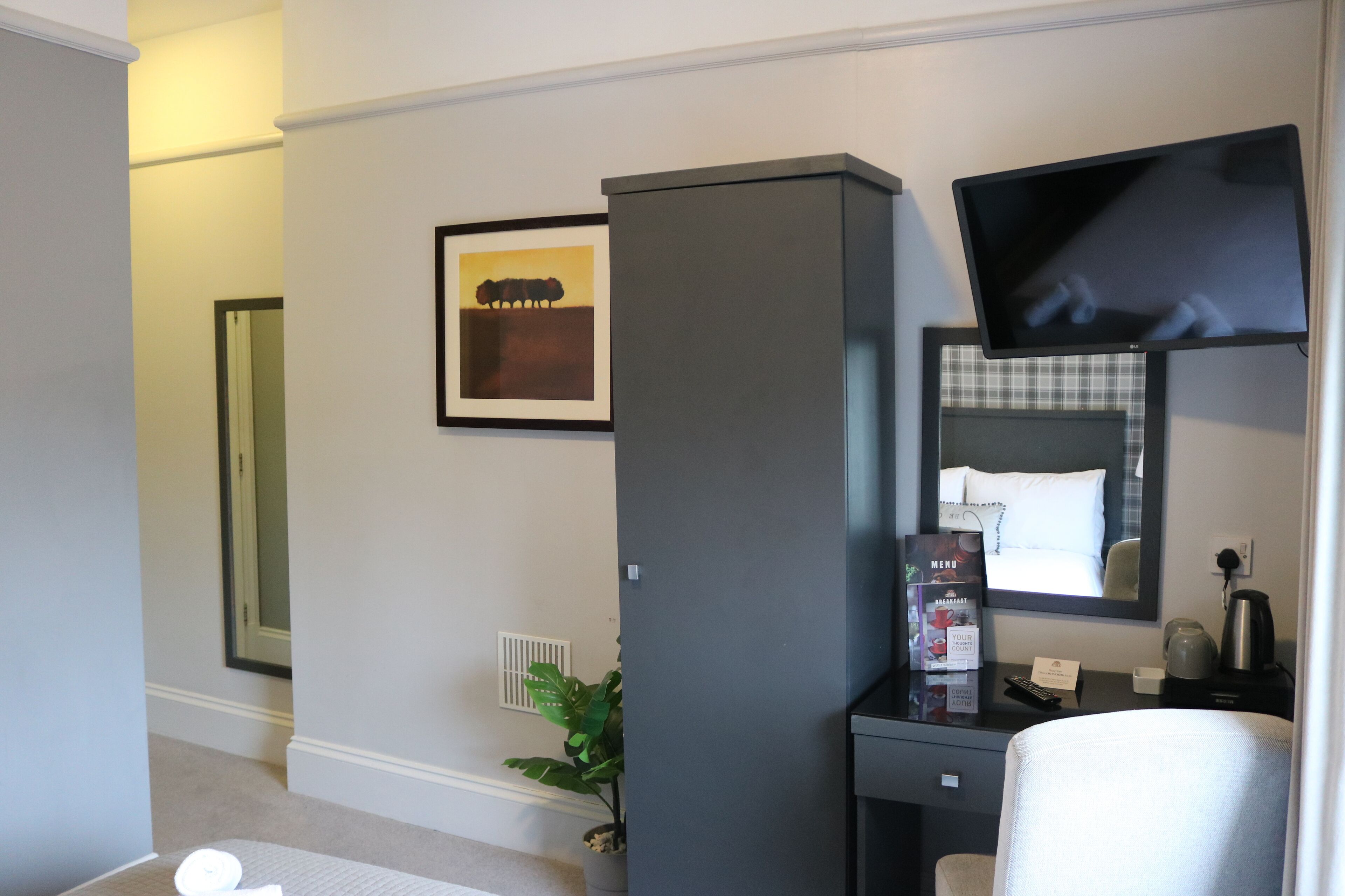 A hotel room with a bed, a desk with a computer, a flat-screen TV, a plant, and a framed picture on the wall.