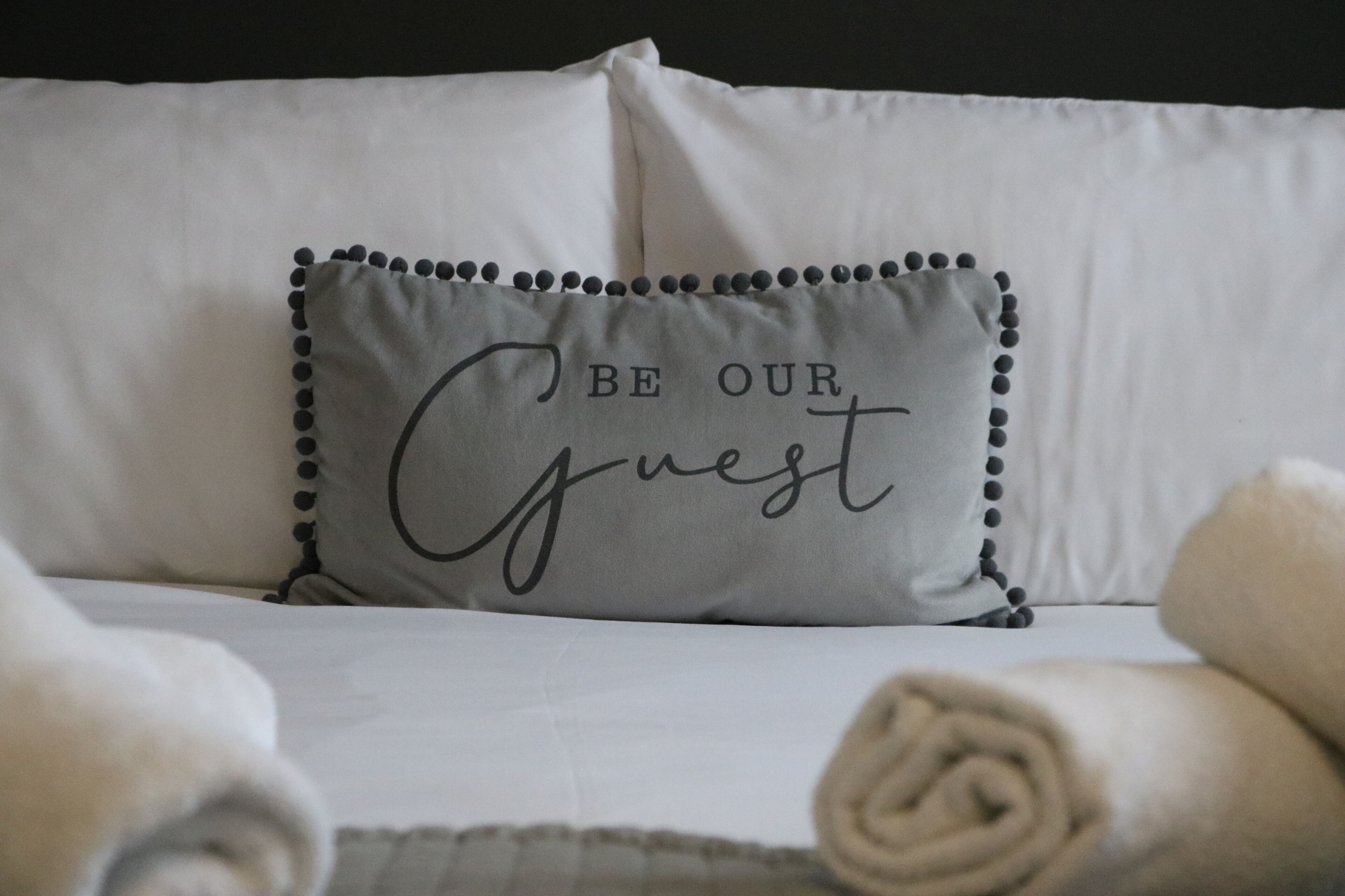 A neatly made bed with a pillow that reads "BE OUR Guest" and rolled towels.