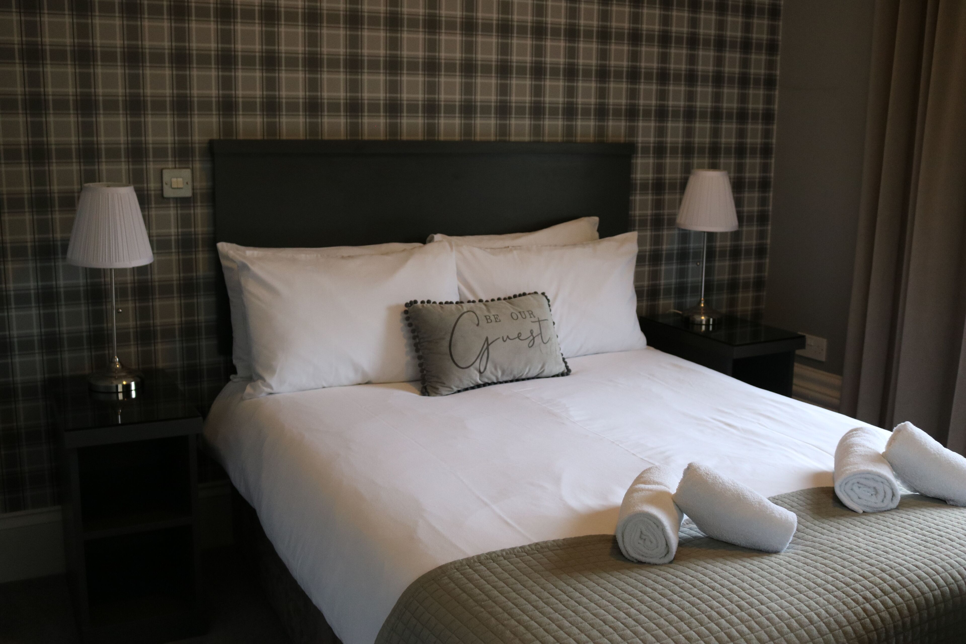 A neatly made bed with white linens, a pillow with a message, and rolled towels on a quilted bedspread.