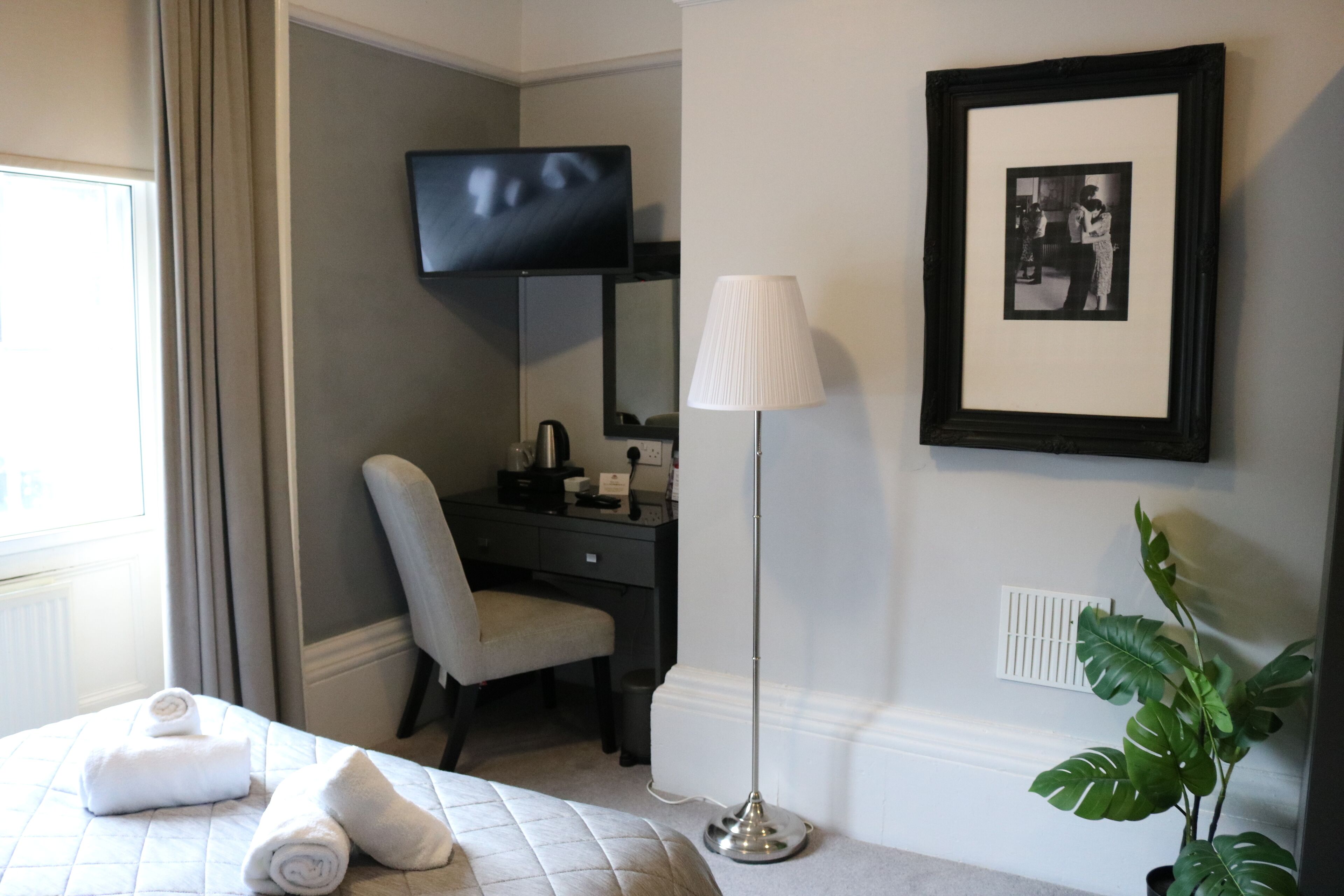 Une chambre d  ’hôtel comprenant un lit, un bureau, une chaise, une télévision, une lampe, une photo encadrée et une plante en pot.
