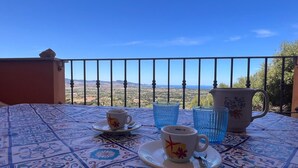 A table with a patterned tablecloth, a cup, a saucer, and a pitcher on a balcony with a clear view of the landscape.