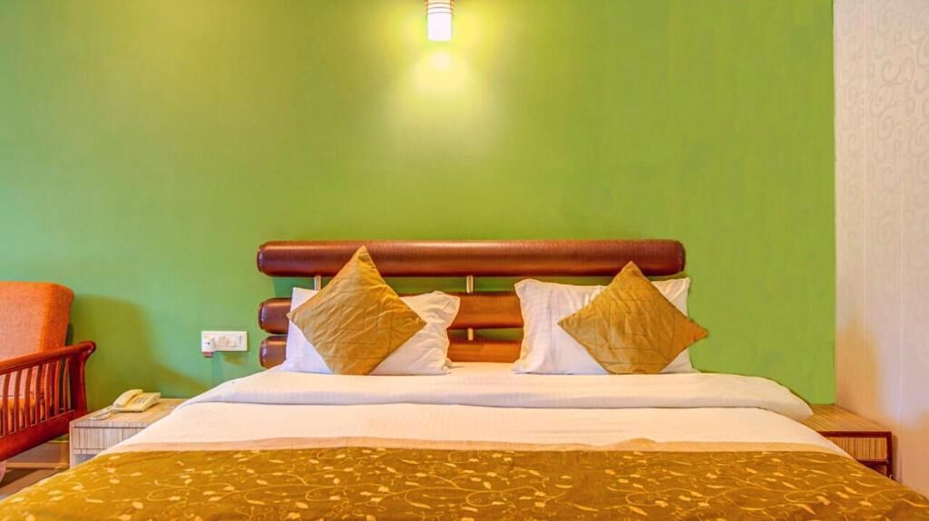 A hotel room with a green wall, a wooden headboard, a bed with white and gold bedding, and a brown throw blanket.