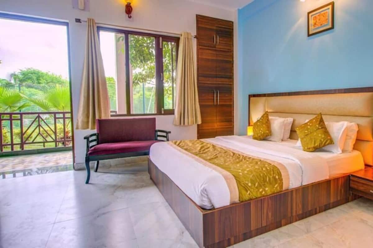 A bedroom with a large bed, a nightstand with a lamp, a window with curtains, and a view of greenery outside.