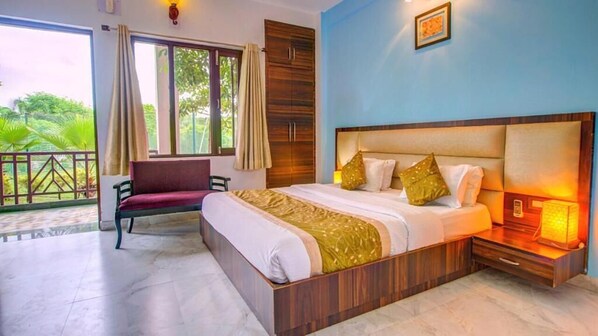 A bedroom with a large bed, a nightstand with a lamp, a window with curtains, and a view of greenery outside. - Pyramid Home Divine (Vikasnagar)