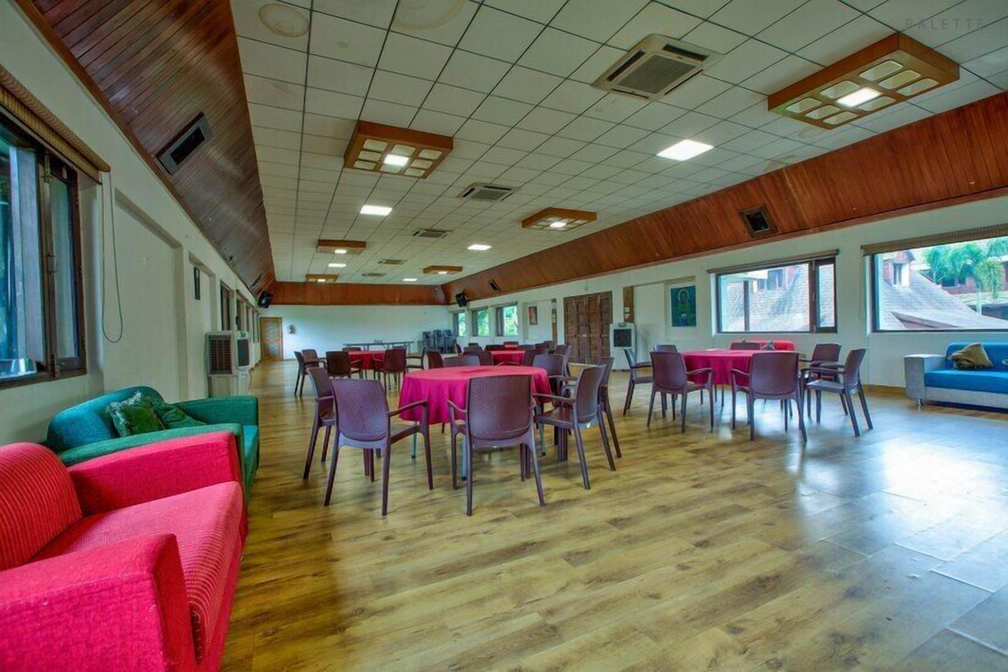 A spacious room with wooden paneling on the ceiling, large windows, and a mix of red and green upholstered seating.