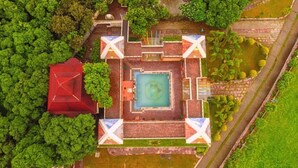 An aerial view of a square building with a central pool, surrounded by greenery and a red-roofed structure.