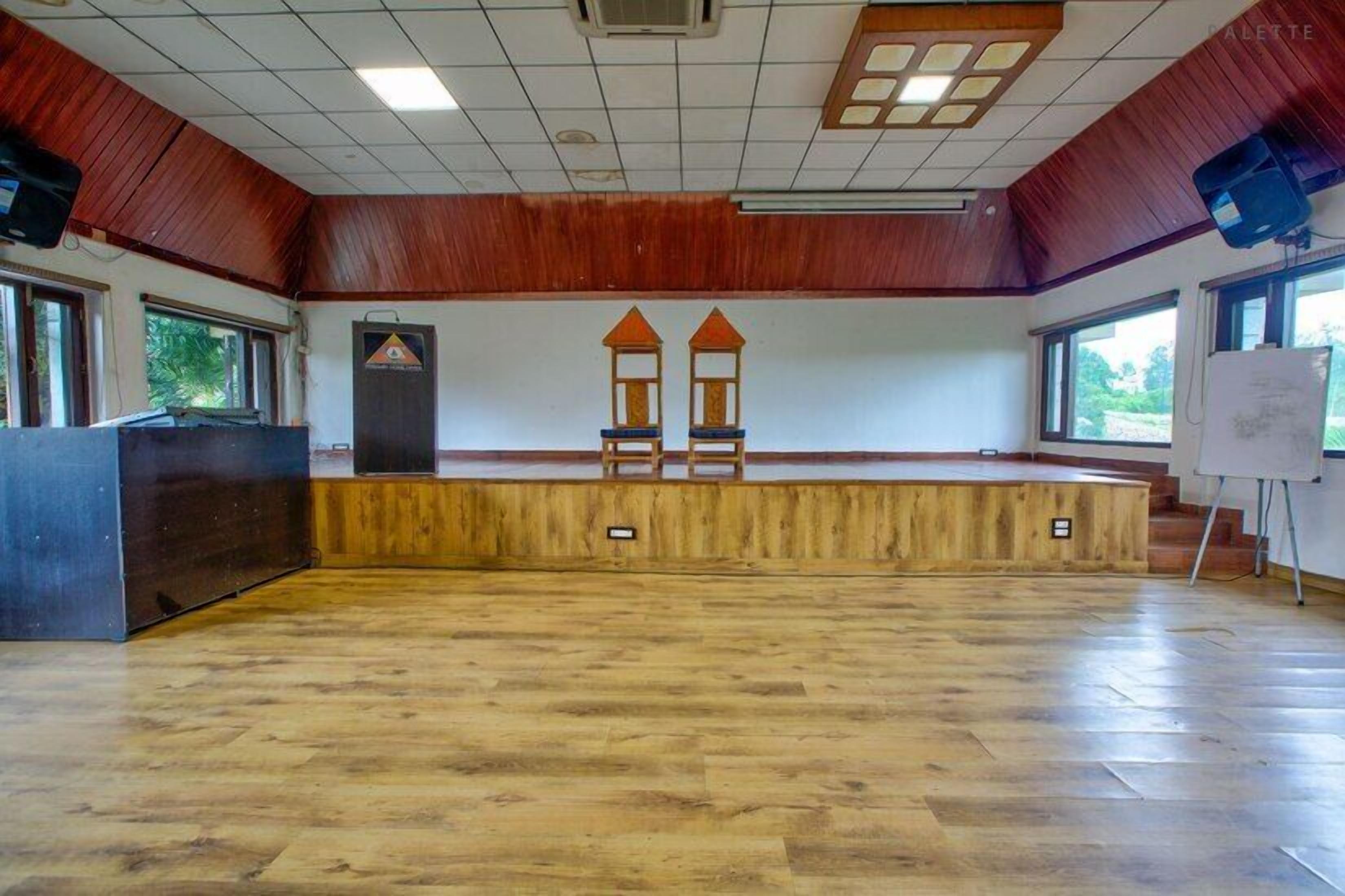 A large hall with a wooden floor, a stage with two chairs, a podium, and a whiteboard.
