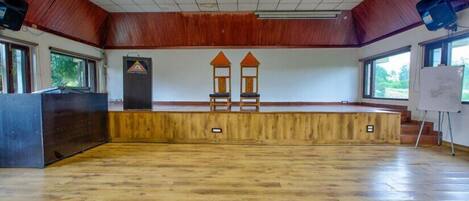 A large hall with a wooden floor, a stage with two chairs, a podium, and a whiteboard.