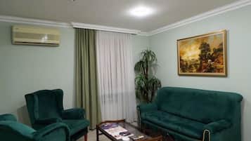 Ein Wohnzimmer mit zwei grünen Sesseln, einem grünen Sofa, einem Holztisch, einem Bild an der Wand, einer Topfpflanze und einer Klimaanlage.