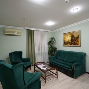 A living room with two green armchairs, a green sofa, a wooden coffee table, a painting on the wall, a potted plant, and an air conditioning unit.