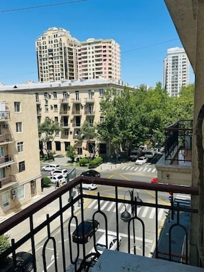 Un balcon donnant sur un immeuble à plusieurs étages, une rue avec des voitures et quelques espaces verts.