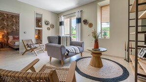 Ein geräumiges Wohnzimmer mit grauem Sofa, Holzsesseln, einem runden Couchtisch und Blick ins Freie durch ein Fenster mit Vorhängen.