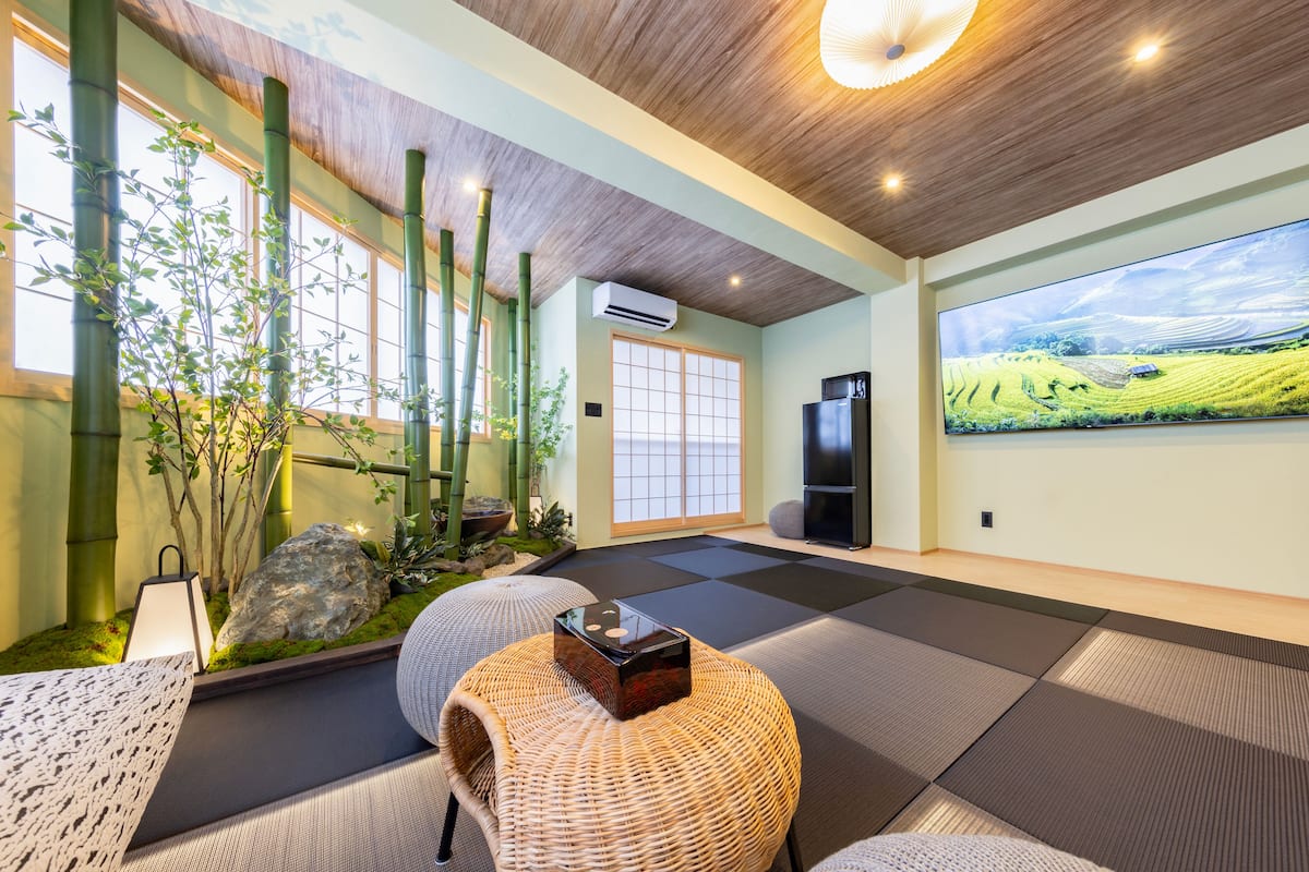 Una habitación moderna con esteras de tatami, un sofá de estilo japonés tradicional, un televisor y un ventanal con vistas a un paisaje verde.