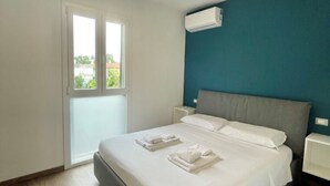 Ein Schlafzimmer mit einem Bett, weißen Leinen, einem grauen Kopfteil, einem Fenster mit Jalousien und einer Klimaanlage an der Wand.