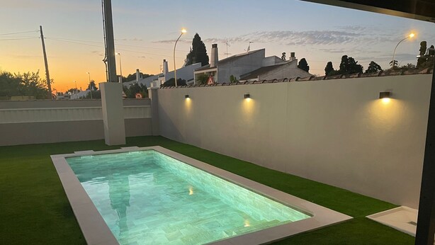 A rectangular swimming pool with a surrounding green artificial turf, set against a backdrop of a sunset sky and residential buildings. - Villa Bolet        . (Chiclana de la Frontera)