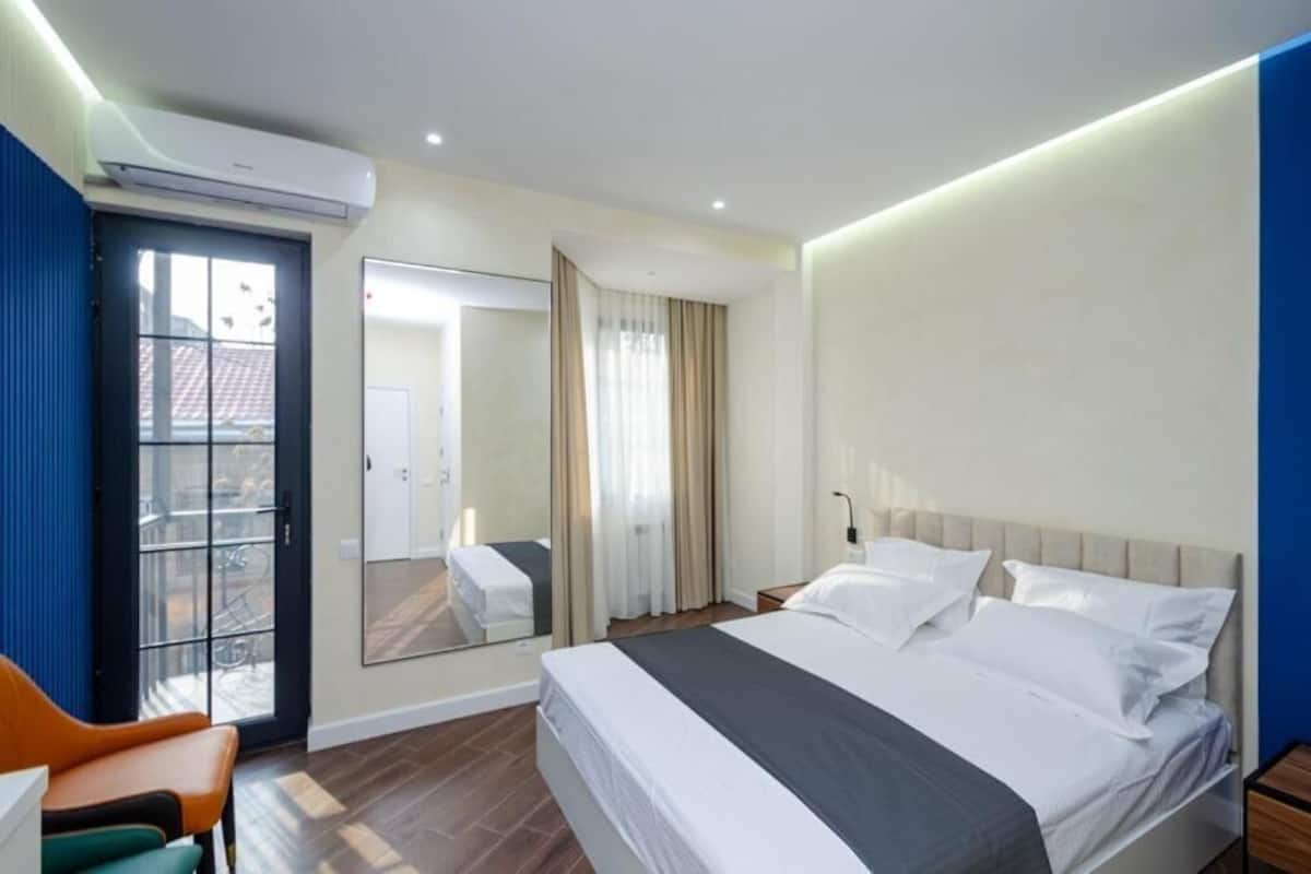 A modern hotel room with a large bed, a seating area with chairs, a mirror, and a view through a glass door.
