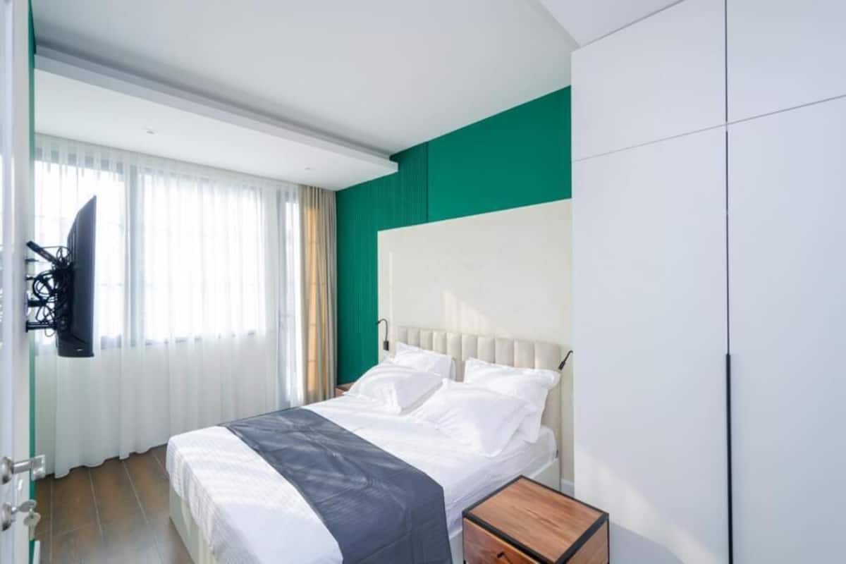 A hotel room with a bed, a nightstand, a TV mounted on the wall, and a window with curtains.
