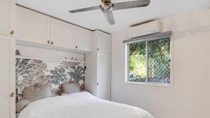 Een slaapkamer met een bed, een plafondventilator, een raam met uitzicht op groen, en witte muren.