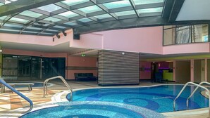 An indoor swimming pool with two hot tubs, a glass ceiling, and a pink wall.