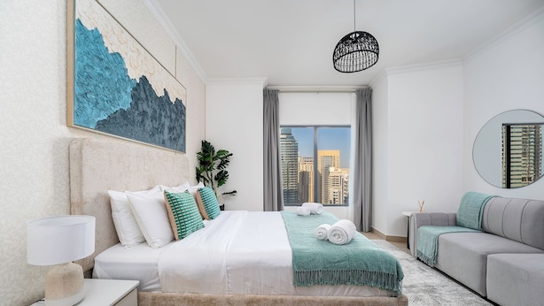 A modern bedroom with a bed, a grey sofa, a nightstand, a lamp, a painting, and a view of the cityscape through the window.