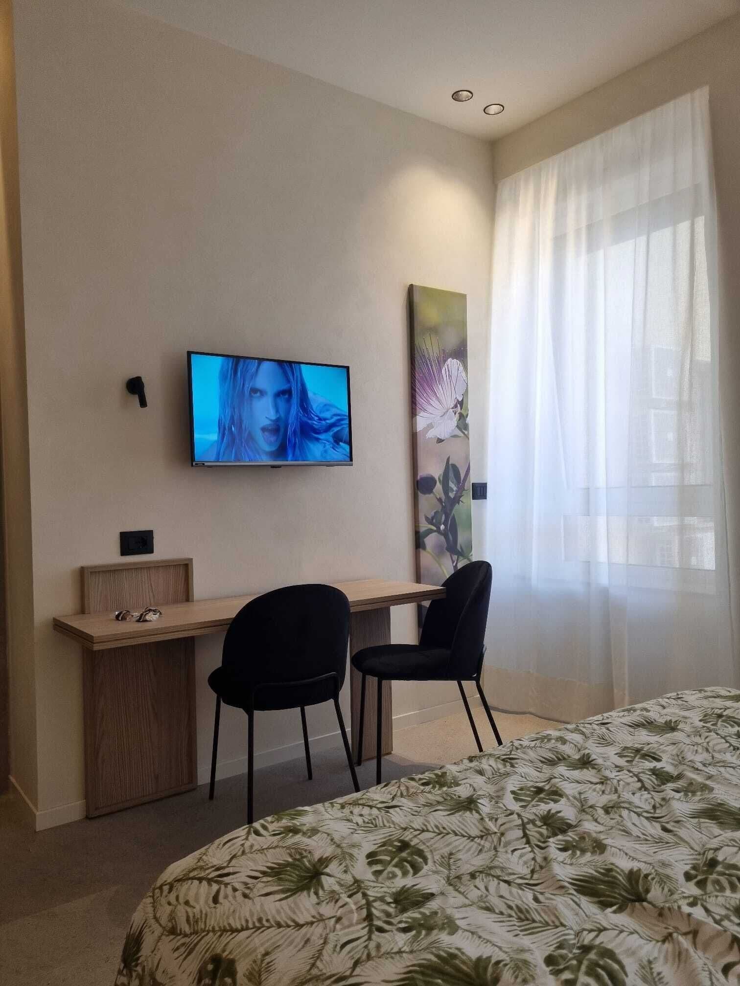 Een hotelkamer met een bed, een bureau met twee stoelen, een aan de muur bevestigde televisie en een groot raam met dunne gordijnen.