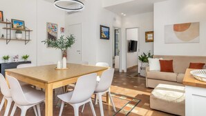 Una sala de estar moderna con una mesa de comedor de madera, sillas blancas, un sofá beige y un televisor montado en la pared.