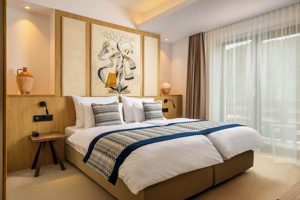 A modern hotel room with a large bed, a nightstand, a wall-mounted lamp, and a large window with sheer curtains.