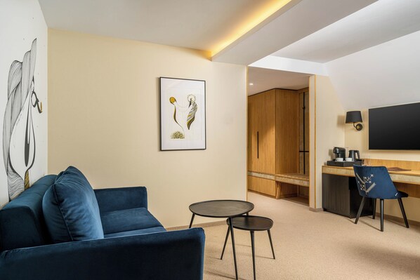 A modern hotel room with a blue sofa, a small round table, a desk with a chair, a TV, and a framed artwork on the wall. - Valedor Hotel Boutique&SPA (Busteni)