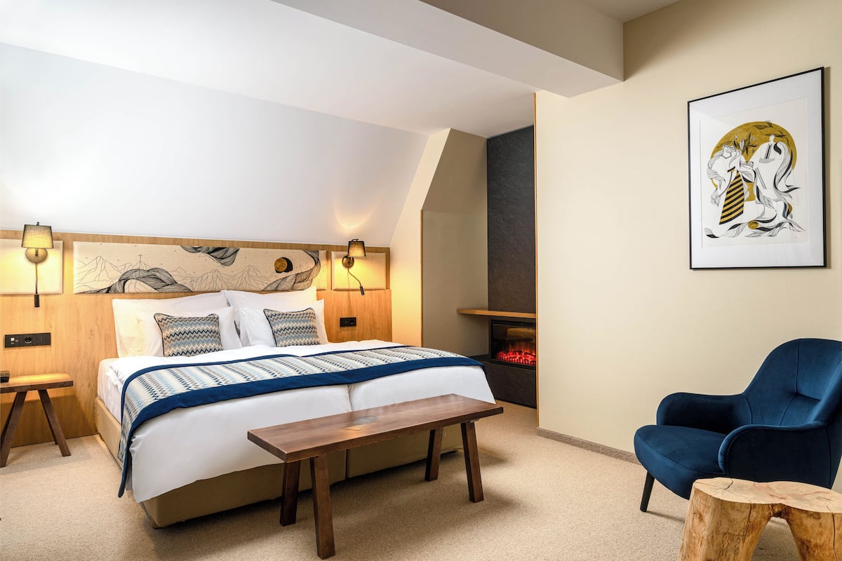 A bedroom with a bed, bedside lamps, a blue chair, a wooden table, and a fireplace.